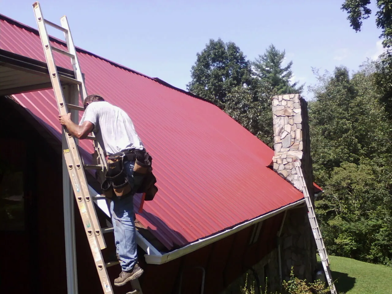 Roofer on a ladder beside a red metal roof and stone chimney — Soffit & Fascia Repair by Roofing Brothers in Morenci